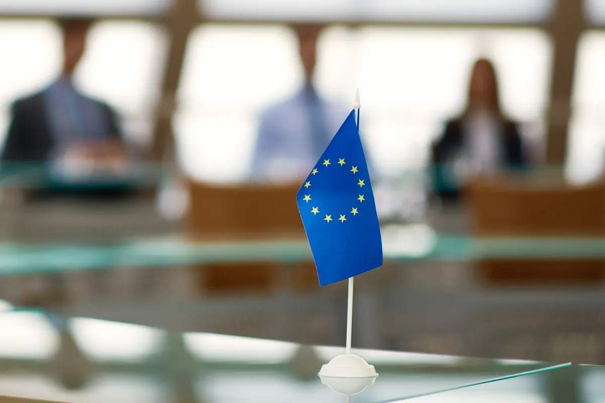 A small European Union flag on a meeting table, with colleagues in conversation blurred behind it